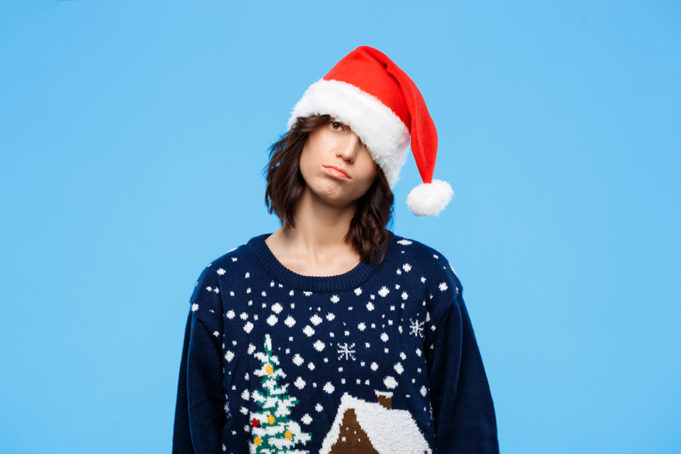 Androgynous person with brown hair wearing an ugly Christmas sweater and Santa hat while frowning against a blue background. - Mila Madison with some tips on how to get through the holiday season as the only transgender person in your family.