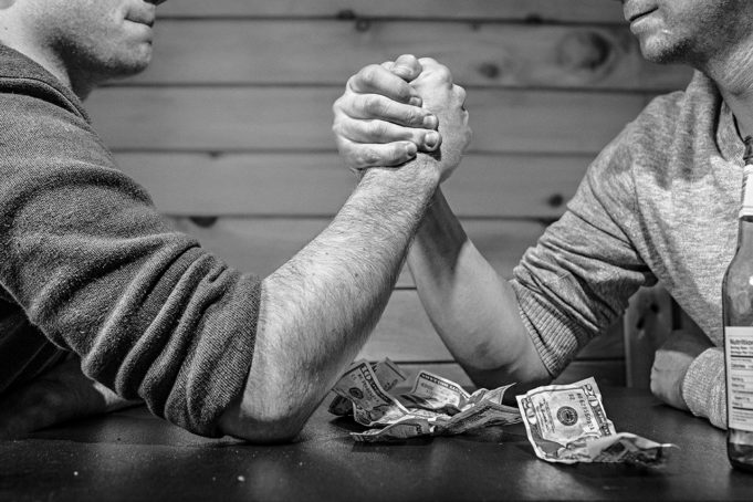 The arms of two men arm wrestling with money on the table - Jude Samson discusses mannerisms and society’s expectation of what a man is, living a stealth lifestyle and how we can change what 'being a man' means.