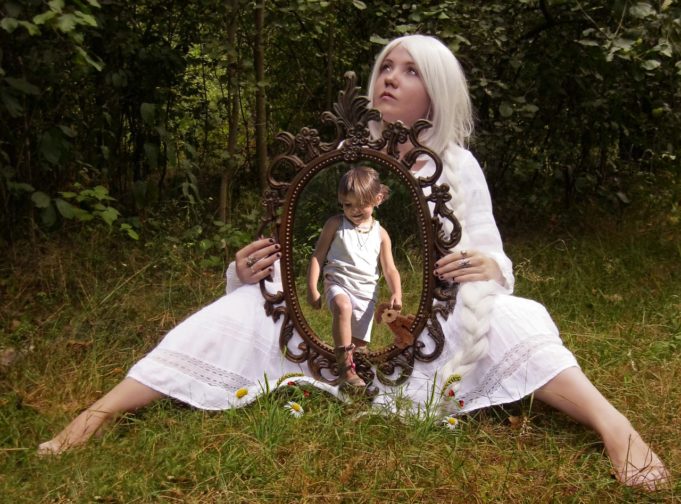 Woman holding a mirror with an image of herself as a child stepping out of it - When a partner’s gender transition and journey to self-acceptance leads to your own realization of who you are. - Trans Partners by U.A. Nigro